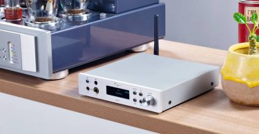 Cayin BTC-2 finition argent posé sur une étagère en bois, écran OLED affichant BT 96KHz LDAC, double sortie casque 6,35 mm et 4,4 mm en façade, antenne Bluetooth déployée, à côté d'un ampli à tubes KT150