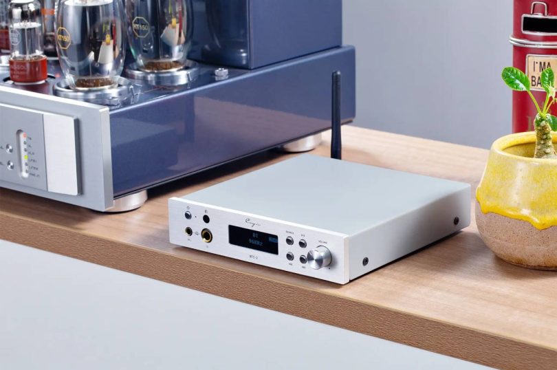 Cayin BTC-2 finition argent posé sur une étagère en bois, écran OLED affichant BT 96KHz LDAC, double sortie casque 6,35 mm et 4,4 mm en façade, antenne Bluetooth déployée, à côté d'un ampli à tubes KT150
