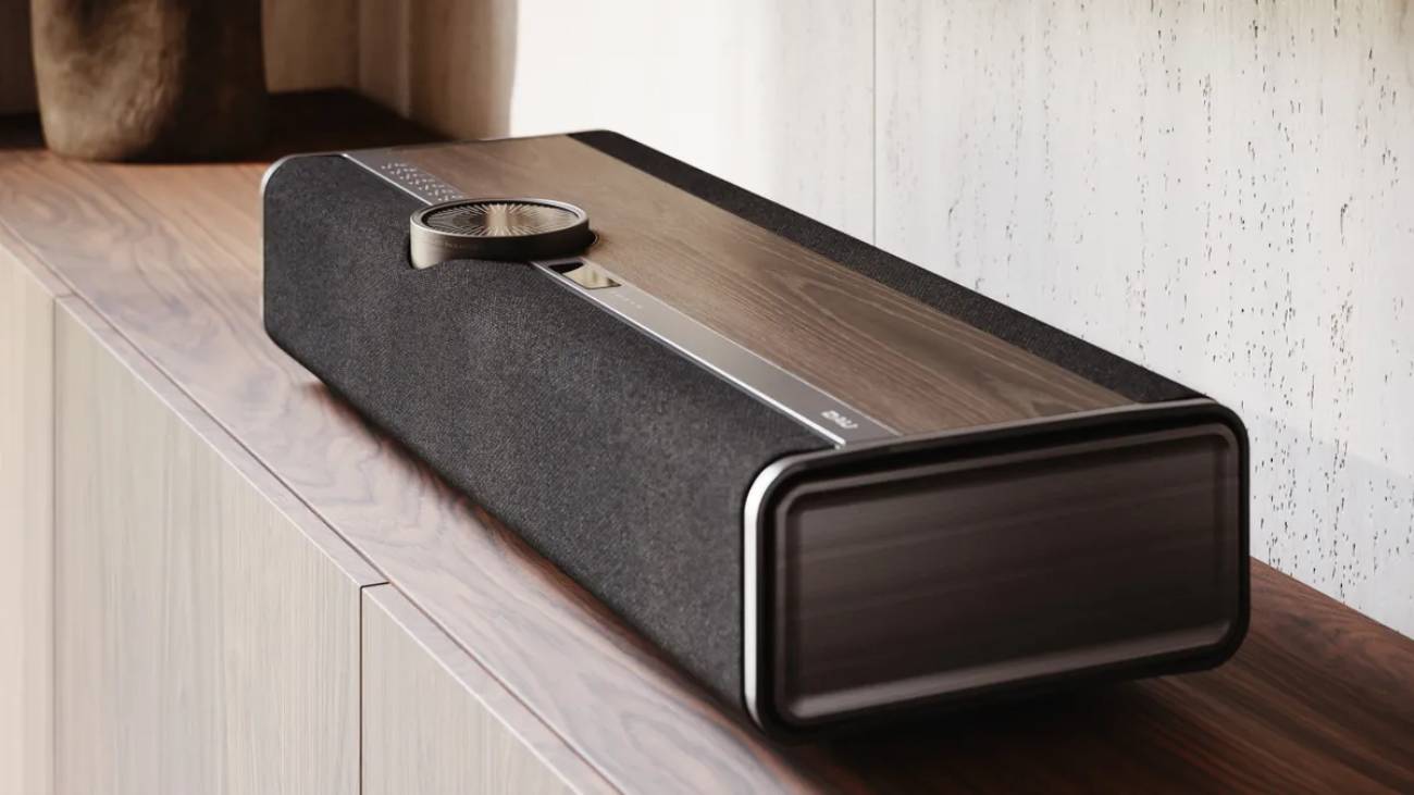 Vue trois-quarts en plongée du système hi-fi tout-en-un DALI VEGA en finition Dark Oak (chêne foncé) posé sur un meuble en noyer, montrant en gros plan la large molette de volume circulaire en aluminium anodisé brossé et verre, l'écran OLED rectangulaire en façade supérieure, le placage bois véritable foncé, la grille en tissu tissé gris anthracite et les coins arrondis du châssis monobloc