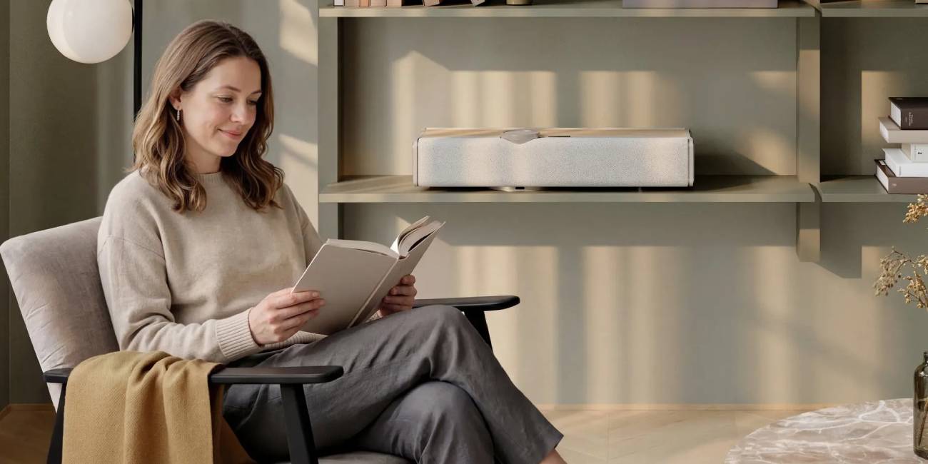 Système hi-fi tout-en-un DALI VEGA en finition Natural Oak posé librement à plat sur une étagère murale en peinture vert sauge dans un intérieur scandinave contemporain, une femme assise dans un fauteuil gris au premier plan lit un livre dans la lumière douce de fin d'après-midi, illustrant le cas d'usage musique d'ambiance pour la lecture et la fonction AOA d'adaptation automatique à l'orientation