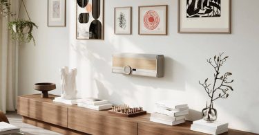 Système hi-fi tout-en-un DALI VEGA en finition Natural Oak fixé au mur en position horizontale dans un salon contemporain scandinave, avec sa large molette de volume centrale en aluminium anodisé et verre, son écran OLED, sa grille tissée et son veneer bois noyer clair, encadré de cadres décoratifs muraux et d'une plante suspendue