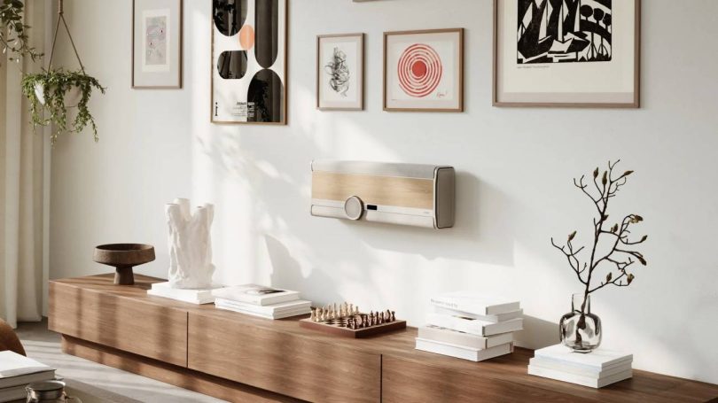 Système hi-fi tout-en-un DALI VEGA en finition Natural Oak fixé au mur en position horizontale dans un salon contemporain scandinave, avec sa large molette de volume centrale en aluminium anodisé et verre, son écran OLED, sa grille tissée et son veneer bois noyer clair, encadré de cadres décoratifs muraux et d'une plante suspendue