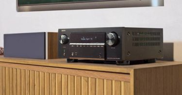 Amplificateur home cinéma Denon AVR-X2900H DAB en finition noire posé sur un meuble bois clair, écran OLED allumé affichant Dolby Surr TV, avec enceinte d'angle grise et tableau encadré au mur en arrière-plan