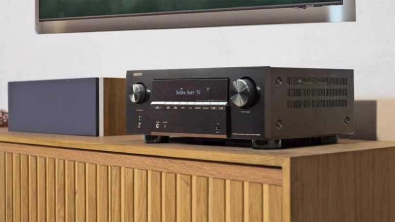Amplificateur home cinéma Denon AVR-X2900H DAB en finition noire posé sur un meuble bois clair, écran OLED allumé affichant Dolby Surr TV, avec enceinte d'angle grise et tableau encadré au mur en arrière-plan