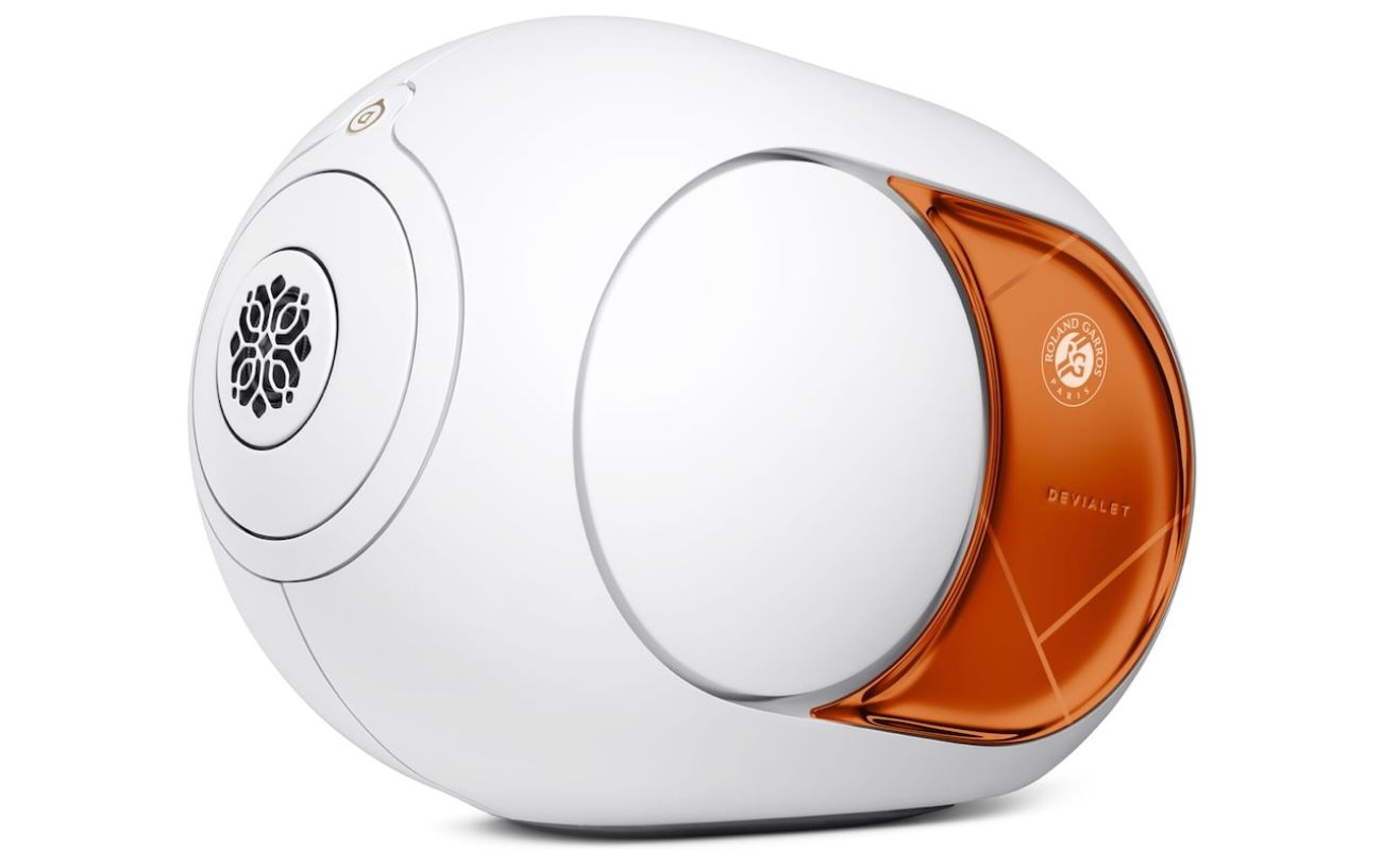 Devialet Phantom Ultimate 108 dB Roland-Garros édition limitée 2026 vue de profil gauche packshot officiel sur fond blanc révélant la silhouette d'œuf caractéristique de la gamme Phantom depuis 2015, corps blanc mat à faible pourcentage de brillance, tweeter à dôme aluminium central blanc émission frontale latérale, woofer arrière en émission latérale opposée à grille décorative ajourée motif floral signature Devialet, flasque latéral droit en finition miroir cuivre orange polissage manuel évoquant la terre battue et la Coupe des Mousquetaires, logo Roland Garros Paris officiel circulaire imprimé en blanc sur le flasque cuivre, signature DEVIALET gravée en relief discret sur le flasque, lignes blanches gravées laser sur le corps blanc évoquant les lignes du court Philippe-Chatrier, bouton tactile cuivre supérieur pour le pilotage local, base socle plate en acier brossé, technologie HBI Heart Bass Implosion descente jusqu'à 14 Hz, 1100 watts d'amplification ADH Analog Digital Hybrid Classe A et Classe D