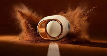 Devialet Phantom Ultimate Roland-Garros Exclusive Edition 2026 vue trois quarts en mise en scène spectaculaire sur terre battue projetée évoquant le court Philippe-Chatrier, finition Clay Red ocre inspirée de la terre battue parisienne, flasque latéral en finition miroir cuivre rose gold figeant le reflet symbolique du court sur l'argent de la Coupe des Mousquetaires, signature DEVIALET gravée discrètement en bas du flasque, logo Roland Garros officiel appliqué en pad printing multicouche autour du dôme central blanc, lignes blanches gravées laser puis vernies haute brillance sur le corps oblong caractéristique de la gamme Phantom depuis 2015, ligne blanche peinte au sol évoquant les lignes du court de tennis, poudre de terre battue projetée autour de l'enceinte rappelant l'énergie cinétique d'une glissade de revers, édition limitée à quelques centaines d'exemplaires disponible dès le 6 mai 2026