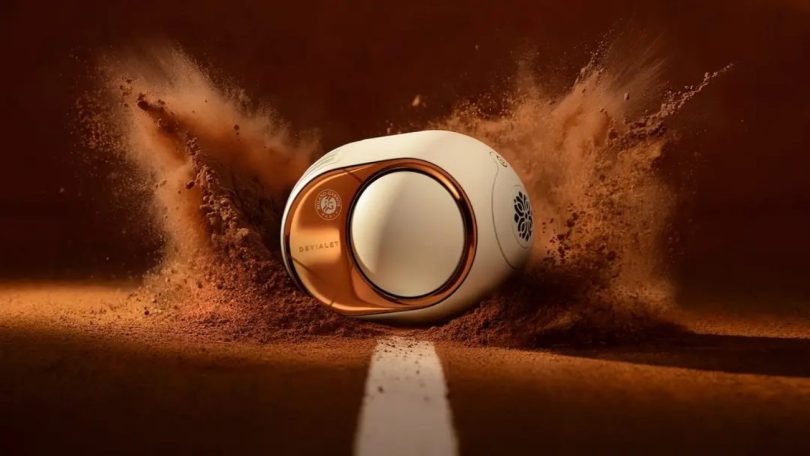 Devialet Phantom Ultimate Roland-Garros Exclusive Edition 2026 vue trois quarts en mise en scène spectaculaire sur terre battue projetée évoquant le court Philippe-Chatrier, finition Clay Red ocre inspirée de la terre battue parisienne, flasque latéral en finition miroir cuivre rose gold figeant le reflet symbolique du court sur l'argent de la Coupe des Mousquetaires, signature DEVIALET gravée discrètement en bas du flasque, logo Roland Garros officiel appliqué en pad printing multicouche autour du dôme central blanc, lignes blanches gravées laser puis vernies haute brillance sur le corps oblong caractéristique de la gamme Phantom depuis 2015, ligne blanche peinte au sol évoquant les lignes du court de tennis, poudre de terre battue projetée autour de l'enceinte rappelant l'énergie cinétique d'une glissade de revers, édition limitée à quelques centaines d'exemplaires disponible dès le 6 mai 2026