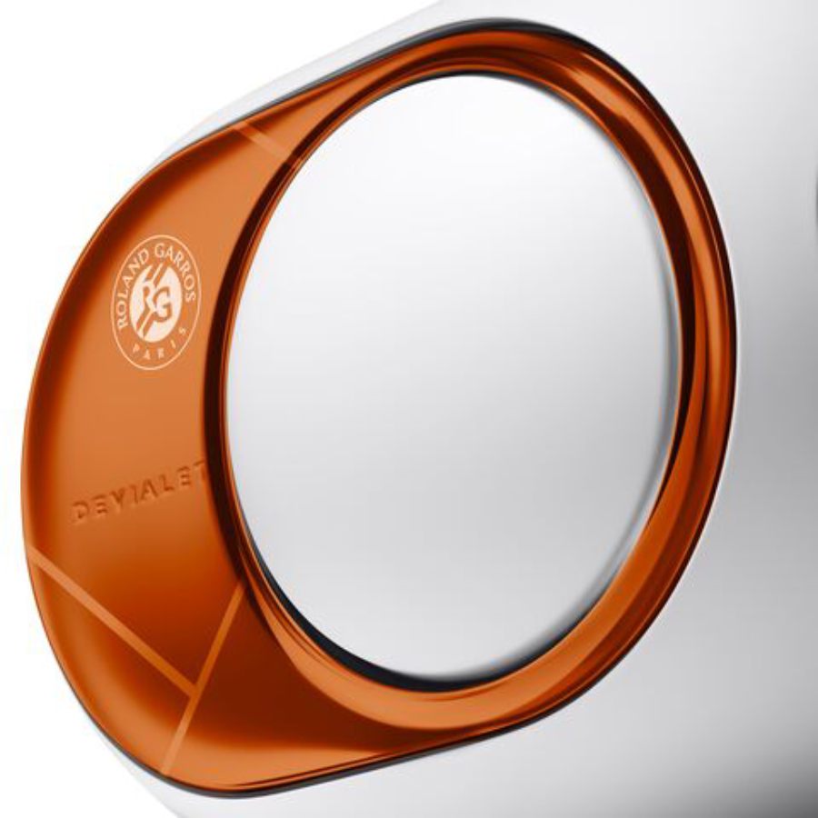 Gros plan macro sur le flasque latéral cuivre orange brillant de la Devialet Phantom Ultimate Roland-Garros édition limitée 2026 montrant le woofer aluminium en émission latérale opposée caractéristique de la technologie HBI Heart Bass Implosion, logo officiel Roland Garros Paris circulaire gravé en blanc dans le coin supérieur gauche du flasque, signature DEVIALET gravée en relief discret en bas du flasque, lignes blanches gravées laser sur le corps blanc de l'enceinte évoquant les lignes du court Philippe-Chatrier, finition miroir poli main sur le flasque cuivre fige le reflet symbolique de la terre battue sur l'argent de la Coupe des Mousquetaires, contraste entre le corps blanc mat à faible pourcentage de brillance et le flasque cuivre haute brillance qui constitue la signature visuelle de cette édition collector limitée à quelques centaines d'exemplaires