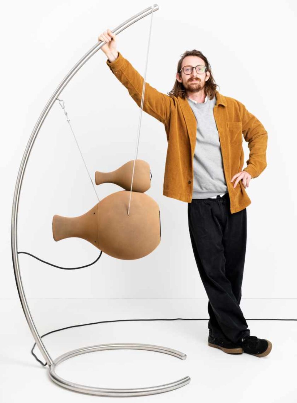 Le designer Anton Erbenich pose à côté de l'enceinte active Econik 1851 suspendue à son arche inox courbée, boîtier bi-sphérique en sable de quartz imprimé 3D ton naturel sable, câbles acier de suspension visibles, photo officielle studio fond blanc