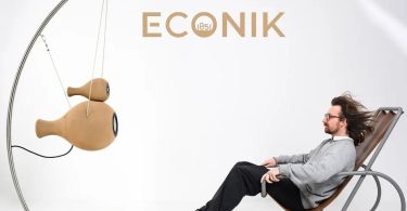 Enceintes actives Econik 1851 en sable de quartz imprimé 3D, suspendues par système de câbles acier sur structure inox courbée, présentées en finition naturelle ton sable à côté d'un auditeur installé sur un fauteuil contemporain, logo ECONIK 1851 au mur en arrière-plan