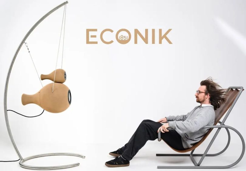 Enceintes actives Econik 1851 en sable de quartz imprimé 3D, suspendues par système de câbles acier sur structure inox courbée, présentées en finition naturelle ton sable à côté d'un auditeur installé sur un fauteuil contemporain, logo ECONIK 1851 au mur en arrière-plan