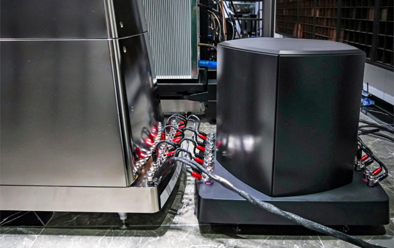 Vue de l'arrière d'une enceinte YG Acoustics Titan posée sur un sol marbré, avec son crossover externe Ultracoherent dans son coffret polymère anti-résonant noir mat positionné à côté, et la batterie de borniers haut-parleur multiples reliés par un câblage cuivre à connecteurs rouge et noir pour multi-amplification