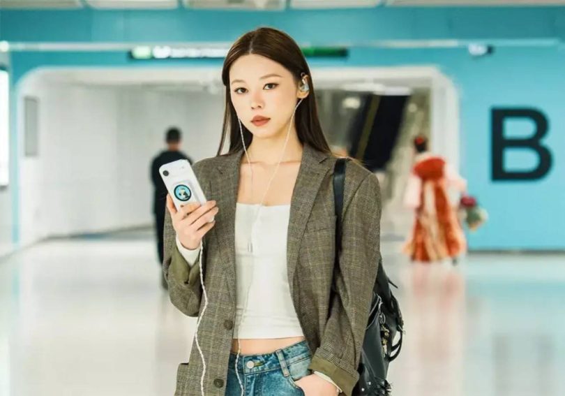 Une utilisatrice en aéroport tient son smartphone équipé du DAC portable Fosi Audio MD3 MagDac fixé magnétiquement au dos, écran circulaire visible, IEM filaires connectés à la sortie casque pour écouter de la musique en haute résolution en déplacement.