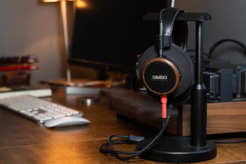 Casque audiophile Grado Signature S550 ouvert avec coque ronde en noyer brésilien massif et logo GRADO blanc, posé sur un support de casque à côté d'un setup de bureau, câble détachable Silver rouge et noir visible