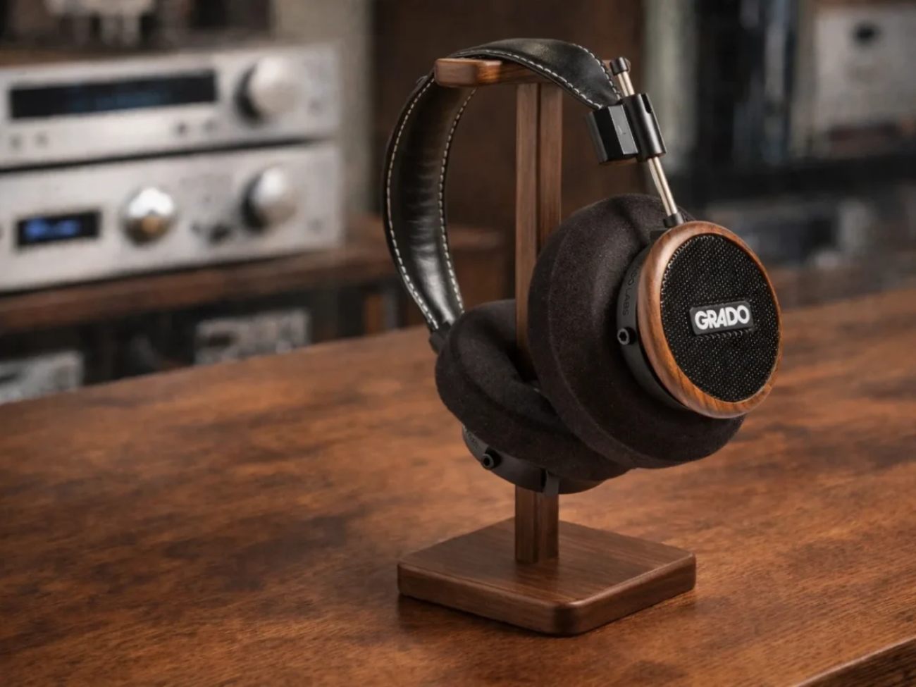 Casque audiophile Grado Signature S550 ouvert posé sur support de casque en bois noyer, coque ronde en noyer brésilien massif visible avec logo GRADO blanc et grille noire, coussinets B-cushion noirs épais, headband cuir noir piqué blanc, en arrière-plan amplis hi-fi vintage flous