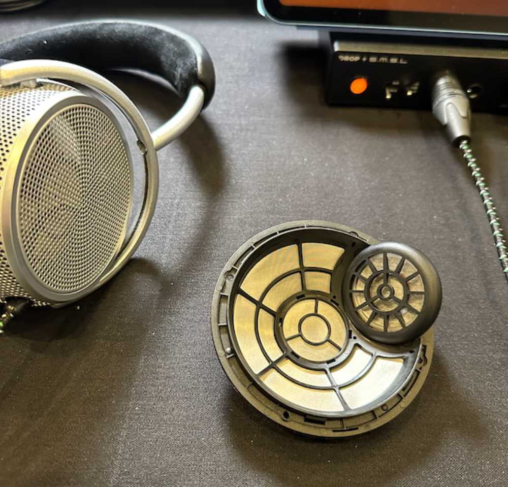 Prototype de driver FSFM Front Side Sound Field Modulation de Grell Audio démonté et exposé sur table, vue en plongée, transducteur principal circulaire de grand diamètre avec structure de guides d'onde en spirale géométrique noire sur membrane argentée, second transducteur secondaire de plus petit diamètre intégré sur la circonférence et orienté en diagonale caractéristique de l'architecture FSFM, contour plastique noir nervuré du châssis du driver, en arrière-plan casque circumaural ouvert Grell Audio avec grille externe à mailles fines et arceau gris argent, amplificateur DAC Drop x S.M.S.L. avec voyant orange allumé, câble symétrique tressé vert et noir connecté en XLR, tablette d'écoute en arrière-plan, ambiance salon audiophile CanJam SoCal 2023