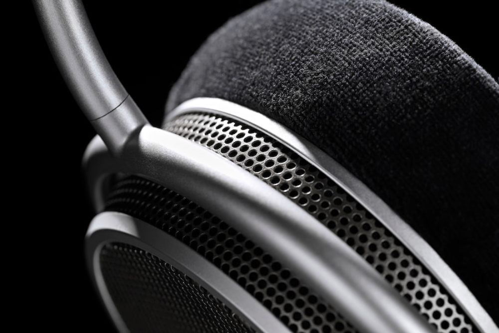 Vue macro en très gros plan du casque Grell Audio OAE2 sur fond noir profond, détail du système de suspension gimbal en aluminium brossé argenté reliant l'arceau à l'écouteur en pivot articulé, coussinet circumaural en velours noir matelassé bien visible à droite, grille de protection externe métallique à perforations hexagonales fines de l'écouteur ouvert visible à gauche et en bas, double épaisseur de grille apparente témoignant de la structure circumaurale ouverte, finition aluminium poli mat de l'arceau et du yoke, ambiance photographique studio haut de gamme