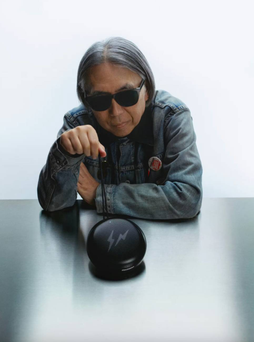 Portrait de Hiroshi Fujiwara fondateur de Fragment Design en veste denim et lunettes noires, accoudé à une table sur laquelle est posée l'enceinte Bang & Olufsen Beosound A1 3rd Gen en édition Fragment, finition aluminium anodisé noir gloss avec logo double éclair visible sous la grille perforée et mention FRAGMENT inscrite sur la tranche