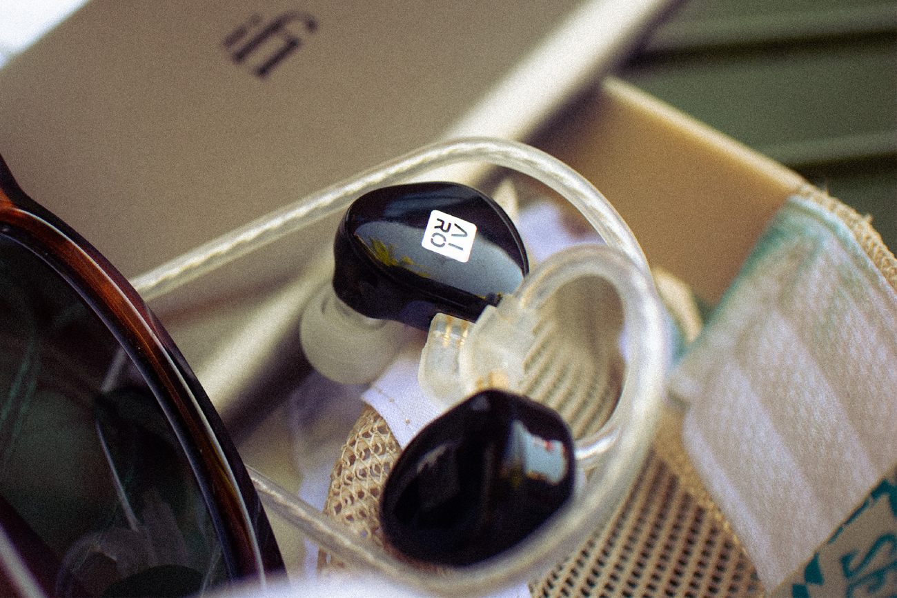 Vue rapprochée d'une coque noire brillante d'écouteur intra-auriculaire iFi AIRO Astra avec logo AIRO visible, ear hook ergonomique transparent et câble 2-pin 0.78 mm transparent. Mise en scène lifestyle estivale avec lunettes de soleil monture écaille, sac en toile et boîte iFi Audio en arrière-plan.