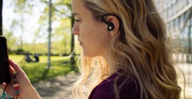 Mise en situation lifestyle d'un écouteur intra-auriculaire iFi Audio AIRO Astra de couleur noire avec ear hook ergonomique, porté par une jeune femme blonde regardant son smartphone dans un parc ensoleillé. Vue de profil montrant le logo iFi visible sur la coque de l'IEM, le câble 2-pin détachable et la forme contour-de-l'oreille.