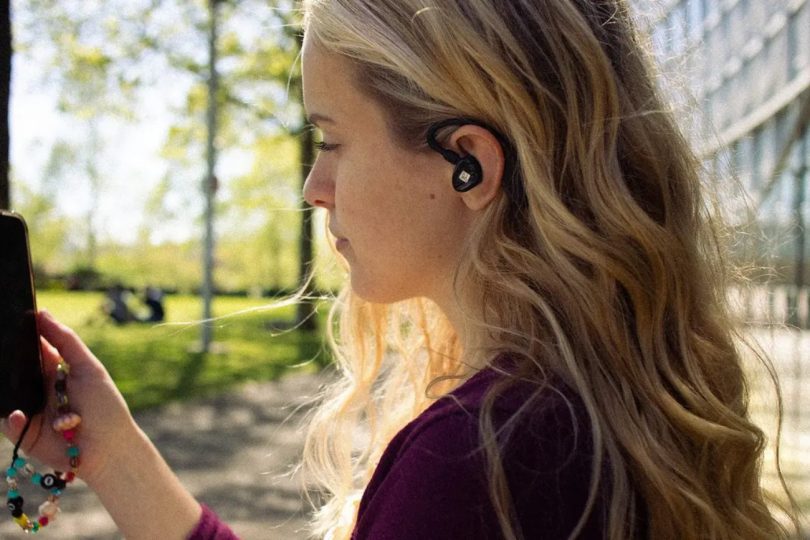 Mise en situation lifestyle d'un écouteur intra-auriculaire iFi Audio AIRO Astra de couleur noire avec ear hook ergonomique, porté par une jeune femme blonde regardant son smartphone dans un parc ensoleillé. Vue de profil montrant le logo iFi visible sur la coque de l'IEM, le câble 2-pin détachable et la forme contour-de-l'oreille.