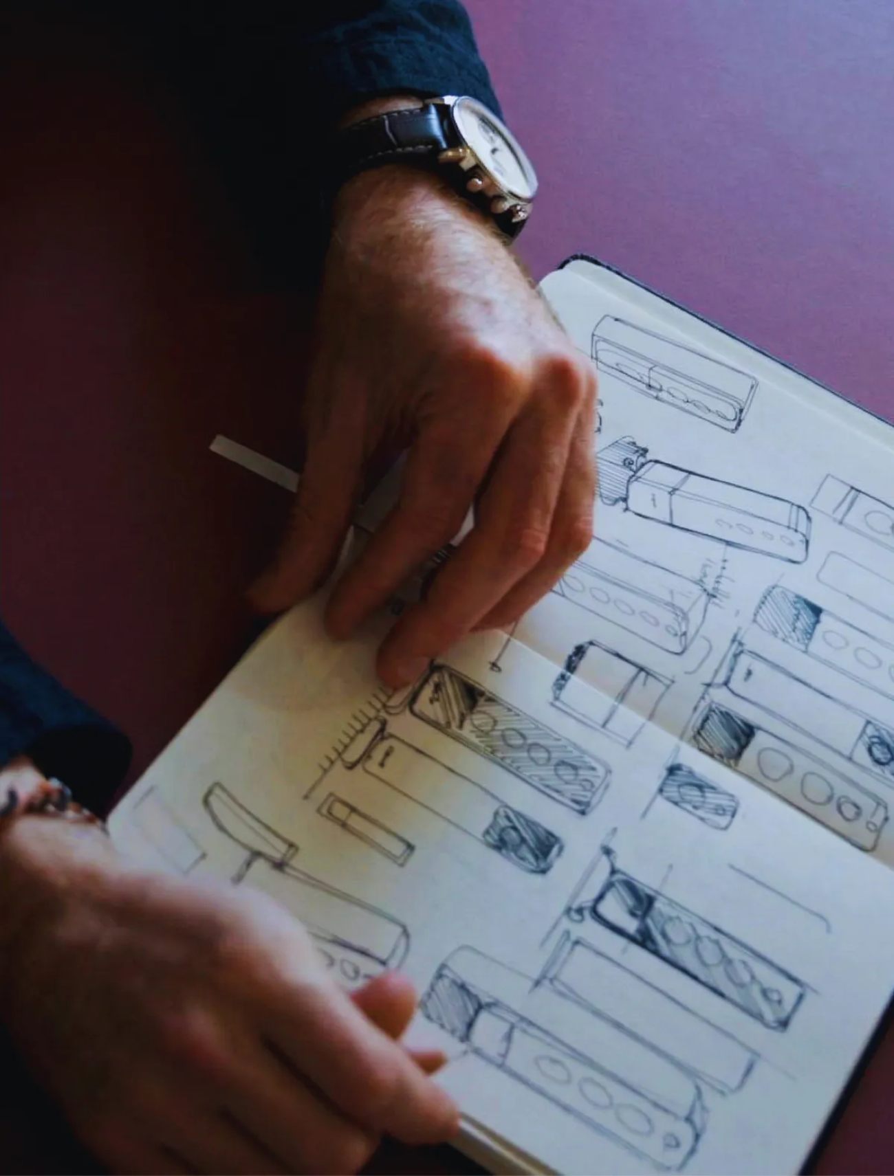 Mains d'un designer industriel feuilletant un carnet de croquis Moleskine rempli d'esquisses préliminaires d'enceintes Jamo au crayon et stylo, processus créatif de l'équipe design pour la nouvelle gamme 2026 incluant des concepts de bookshelf colonne soundbar et satellites multi-haut-parleurs en exploration scandinave