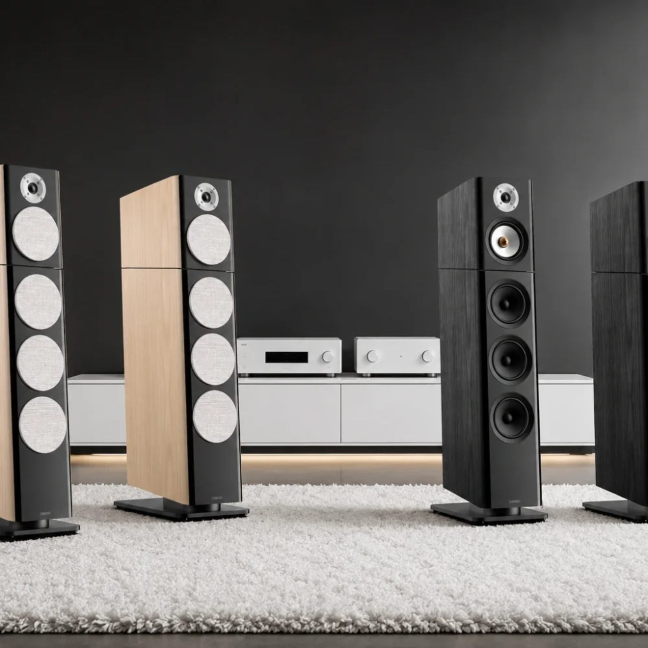 Comparatif visuel de quatre enceintes colonnes haut de gamme alignées sur un tapis blanc dans un salon contemporain, paire Jamo Concert Legacy en finitions Onyx noire et Heritage chêne clair à gauche avec leurs grilles tissu rondes signature, paire d'enceintes colonnes concurrentes au tweeter dôme apparent et triple woofer à cônes noirs visibles à droite, meuble bas blanc rétroéclairé avec amplification hi-fi argentée et lecteur réseau au centre