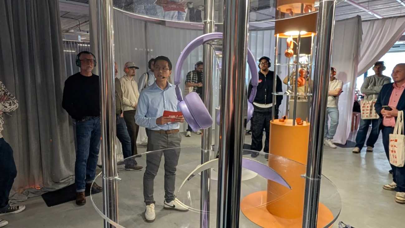 Présentation officielle des nouveaux casques JBL Live 780NC en coloris violet (Purple) et orange à l'événement Club JBL Amsterdam pour le 80ème anniversaire de la marque, avec présentoirs cylindriques en plexiglas et public en écoute sur casques