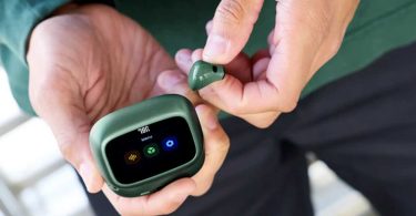 Vue rapprochée du boîtier de charge Smart Charging Case de la gamme JBL Live 4 en finition vert sapin, écran tactile allumé affichant le menu principal avec icônes équaliseur, logo JBL et paramètres, écouteur compact assorti tenu entre les doigts d'un utilisateur