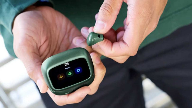 Vue rapprochée du boîtier de charge Smart Charging Case de la gamme JBL Live 4 en finition vert sapin, écran tactile allumé affichant le menu principal avec icônes équaliseur, logo JBL et paramètres, écouteur compact assorti tenu entre les doigts d'un utilisateur