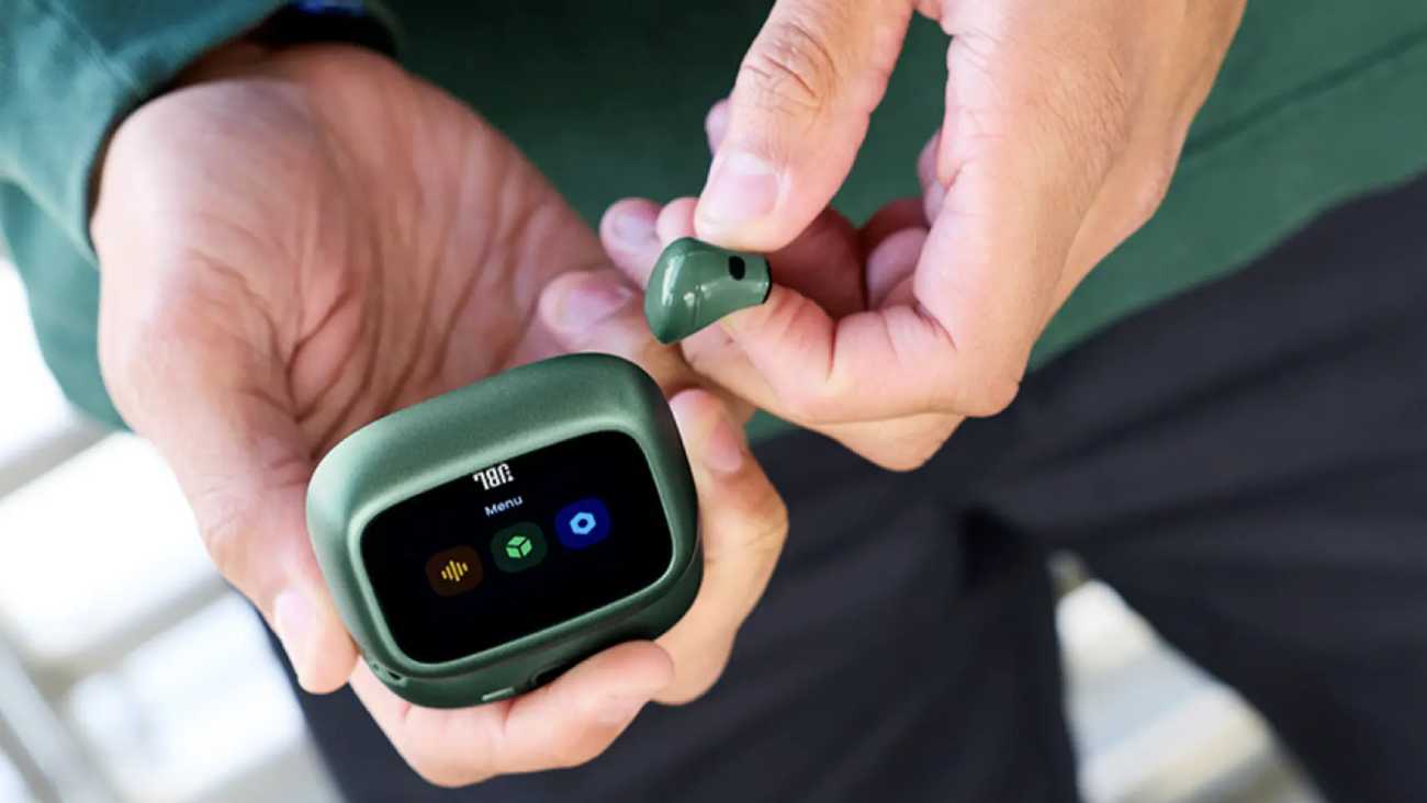 Vue rapprochée du boîtier de charge Smart Charging Case de la gamme JBL Live 4 en finition vert sapin, écran tactile allumé affichant le menu principal avec icônes équaliseur, logo JBL et paramètres, écouteur compact assorti tenu entre les doigts d'un utilisateur