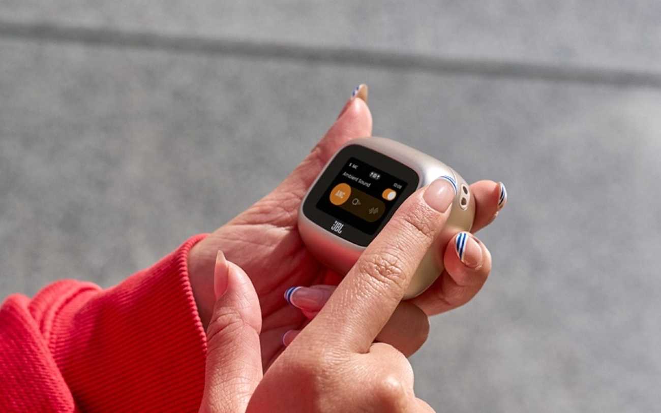 Boîtier de charge intelligent Smart Charging Case de la gamme JBL Live 4 en finition sable tenu entre les mains d'une utilisatrice, écran tactile allumé affichant l'interface Ambient Sound avec sélecteurs de réduction de bruit active ANC, mode neutre et mode transparence, manche de sweat rouge visible en arrière-plan