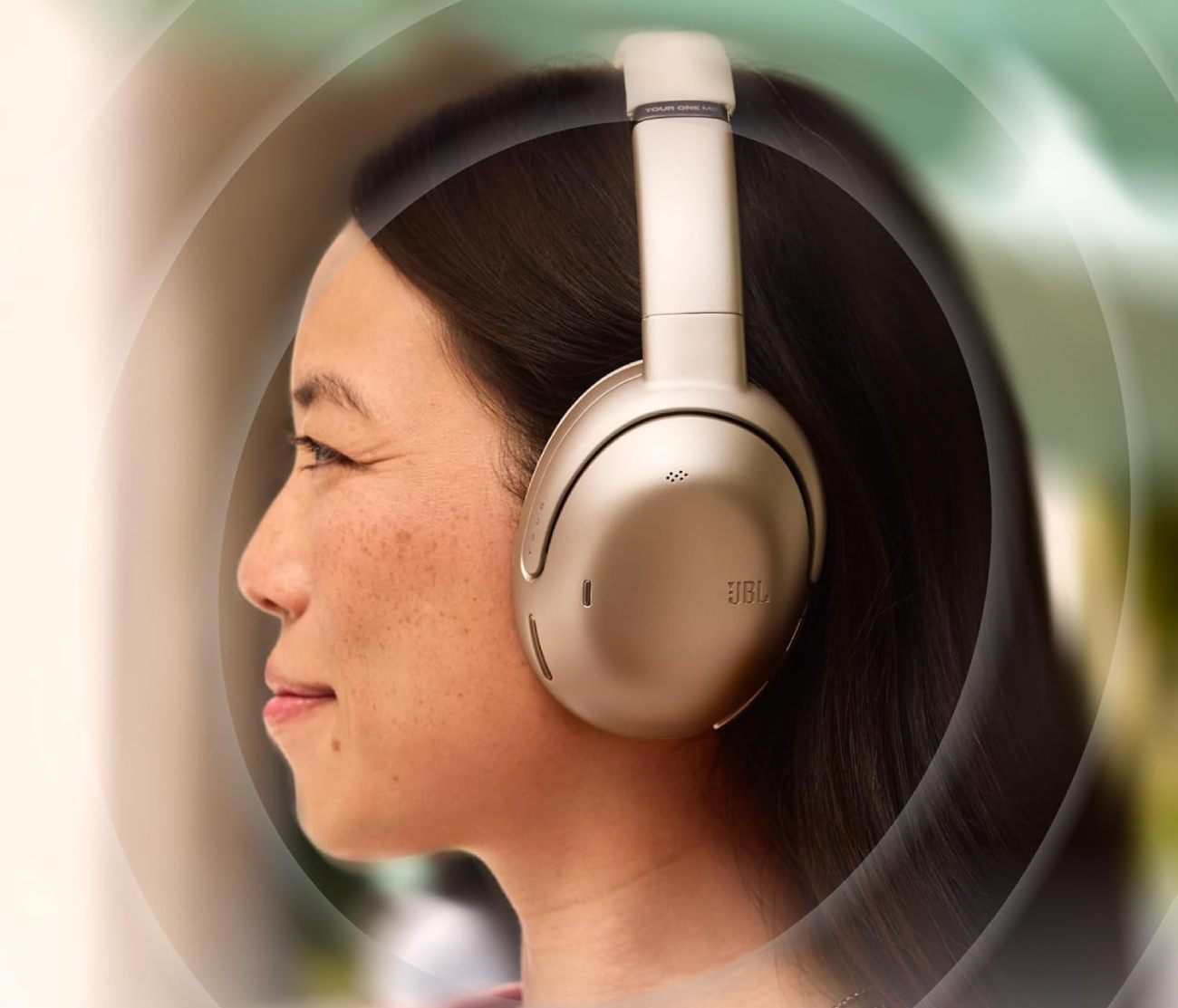 Femme aux cheveux noirs portant le casque circum-aural sans fil JBL Tour ONE M3 dans le coloris champagne mocha crème, vue de profil de trois-quarts gauche, oreillette circulaire ergonomique avec marquage TOUR et logo JBL, microphone ANC visible sur le cache extérieur, arceau ajustable avec gravure TOUR ONE M3