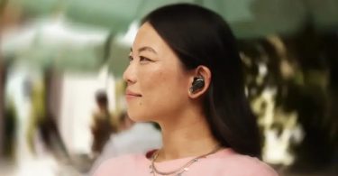 Femme aux cheveux noirs longs portant des écouteurs True Wireless JBL Tour PRO 3 noirs dans l'oreille, vue de profil de trois-quarts dans un environnement urbain extérieur ensoleillé en arrière-plan flou, ambiance lifestyle illustrant l'usage nomade des écouteurs avec mise à jour firmware Sound Curve 2026