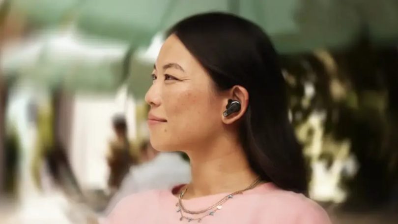 Femme aux cheveux noirs longs portant des écouteurs True Wireless JBL Tour PRO 3 noirs dans l'oreille, vue de profil de trois-quarts dans un environnement urbain extérieur ensoleillé en arrière-plan flou, ambiance lifestyle illustrant l'usage nomade des écouteurs avec mise à jour firmware Sound Curve 2026
