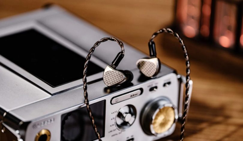 Paire d'IEMs Kiwi Ears Halcyon en finition métal argent avec faceplate à motif répété KE et câble OFC tressé 2-pin, posée sur un DAP audiophile haut de gamme avec sorties multiples 4.4mm, 3.5mm, 6.35mm et XLR4
