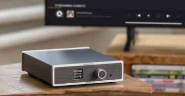 Streaming transport Lindemann Woodnote Bridge en chassis aluminium argent et anthracite avec façade boisée arborant les marquages WOOD BRIDGE NOTE et un large bouton de commande, posé sur table en bois clair devant un écran de TV affichant une interface de streaming musical en arrière-plan flou