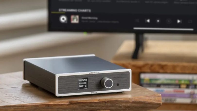 Streaming transport Lindemann Woodnote Bridge en chassis aluminium argent et anthracite avec façade boisée arborant les marquages WOOD BRIDGE NOTE et un large bouton de commande, posé sur table en bois clair devant un écran de TV affichant une interface de streaming musical en arrière-plan flou