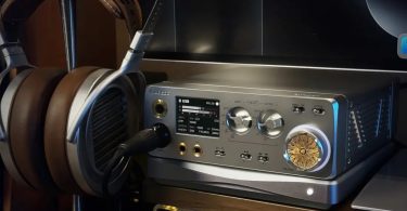 Vue frontale du Lotoo Gungnir DAC ampli casque de bureau en situation d'écoute, avec casque planar magnétique HiFiMan posé sur support à gauche et câble symétrique 4.4mm Pentaconn branché sur la sortie casque, écran TFT central affichant le mode USB volume 50 entrée LTTP format DSD 176.4 kHz 24 bits gain -10 dB sortie 2 Vrms, deux molettes argentées de sélection INPUT et OUTPUT avec positions LTTP USB OPT COAX AES et 4.4/3.5 LO DO, commutateurs MENU OK et POWER, boutons F1 F2 Play Pause piste précédente suivante, imposante molette de volume dorée gravée du logo Lotoo Helios entourée d'un anneau LED bleu, châssis aluminium gris brossé avec dissipateur thermique latéral à ailettes, étagère bois naturel