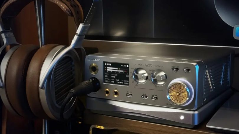 Vue frontale du Lotoo Gungnir DAC ampli casque de bureau en situation d'écoute, avec casque planar magnétique HiFiMan posé sur support à gauche et câble symétrique 4.4mm Pentaconn branché sur la sortie casque, écran TFT central affichant le mode USB volume 50 entrée LTTP format DSD 176.4 kHz 24 bits gain -10 dB sortie 2 Vrms, deux molettes argentées de sélection INPUT et OUTPUT avec positions LTTP USB OPT COAX AES et 4.4/3.5 LO DO, commutateurs MENU OK et POWER, boutons F1 F2 Play Pause piste précédente suivante, imposante molette de volume dorée gravée du logo Lotoo Helios entourée d'un anneau LED bleu, châssis aluminium gris brossé avec dissipateur thermique latéral à ailettes, étagère bois naturel