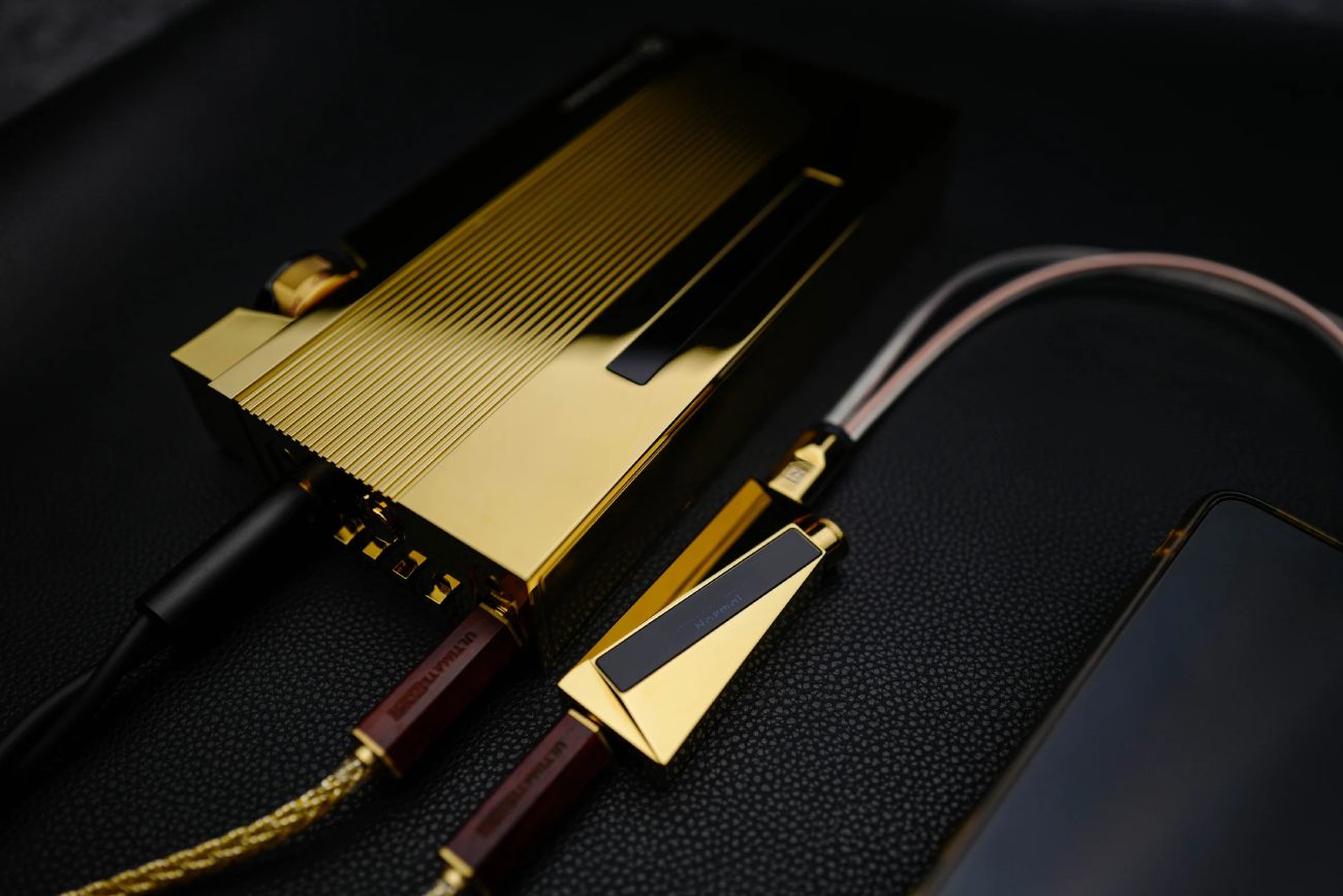 Mise en scène du Luxury & Precision W4 ACG Mirror Copper Edition (premier plan, dongle doré avec écran OLED) connecté en line-out fixed voltage à l'ampli portable EA4 Mirror Copper Edition (grand boîtier doré aux ailettes verticales) via le câble Y WY1 Mirror Copper, avec smartphone et IEM audiophile sur surface en cuir noir grené