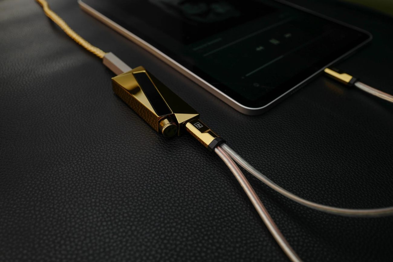 Vue rapprochée du dongle DAC Luxury & Precision W4 ACG Mirror Copper Edition en situation d'écoute, connecté en USB-C à une tablette tactile via le câble Y WY1 Mirror Copper Special Edition à gaine tressée dorée, sortie casque 4.4 mm balanced reliée à un câble d'IEM transparent multibrins, molette ALPS de contrôle visible, le tout posé sur surface en cuir noir grené