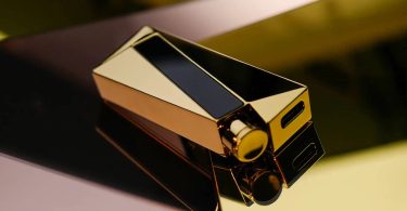 Dongle DAC portable Luxury & Precision W4 ACG Mirror Copper Edition vu en plongée trois quarts, boîtier en cuivre poli miroir avec revêtement nitrure de titane doré, écran OLED noir intégré sur la face supérieure, port USB-C visible sur le côté droit, posé sur une surface réfléchissante avec jeu de lumière dorée