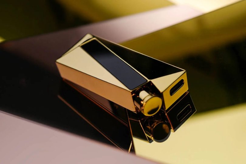 Dongle DAC portable Luxury & Precision W4 ACG Mirror Copper Edition vu en plongée trois quarts, boîtier en cuivre poli miroir avec revêtement nitrure de titane doré, écran OLED noir intégré sur la face supérieure, port USB-C visible sur le côté droit, posé sur une surface réfléchissante avec jeu de lumière dorée