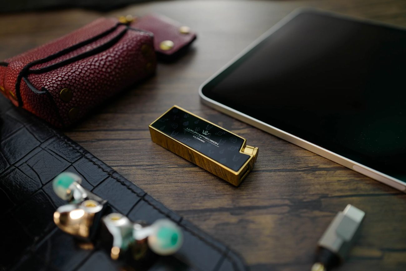 Mise en scène lifestyle du Luxury & Precision W4 ACG Mirror Copper Edition posé sur une table en bois sombre veiné, écran OLED allumé affichant le logo Luxury & Precision et le mode Classic avec icônes culturelles ACG en filigrane, entouré d'un étui en cuir galuchat rouge bordeaux à fermeture aimantée, d'une tablette tactile, d'IEMs audiophiles à coque transparente verte avec connecteurs MMCX cuivre et d'un adaptateur USB-C