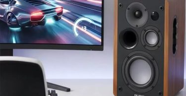 Enceinte Majority D100X en finition noyer Walnut avec 3 drivers visibles (tweeter, médium, woofer) et port basse-réflex frontal, posée sur un bureau à côté d'un écran d'ordinateur affichant un jeu de course automobile