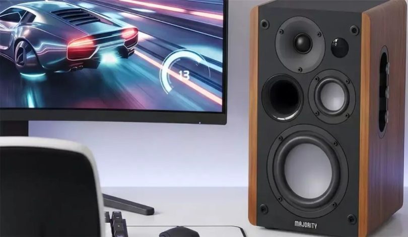Enceinte Majority D100X en finition noyer Walnut avec 3 drivers visibles (tweeter, médium, woofer) et port basse-réflex frontal, posée sur un bureau à côté d'un écran d'ordinateur affichant un jeu de course automobile