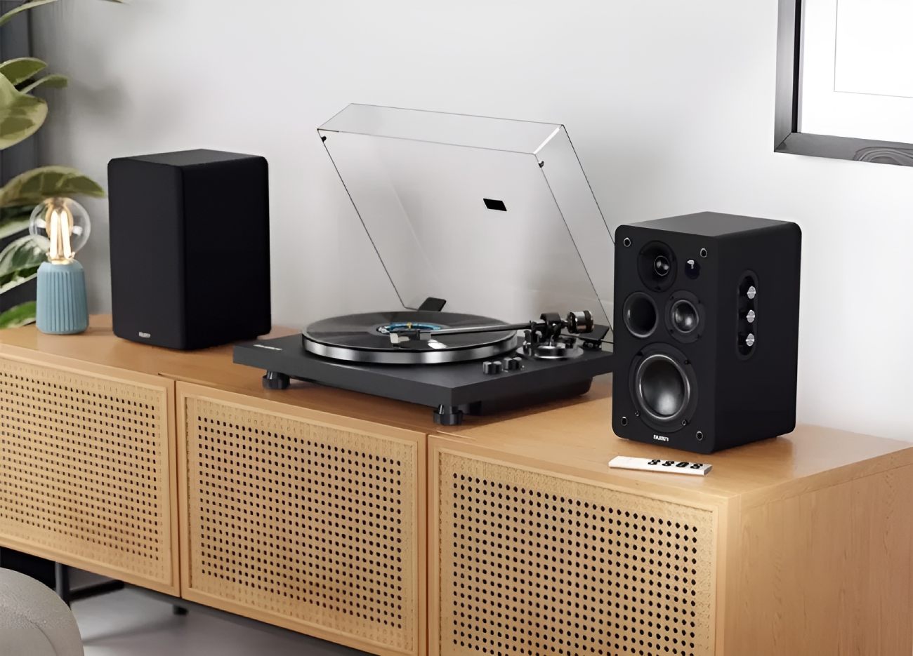 Paire d'enceintes Majority D100X en finition noir laqué encadrant une platine vinyle posée sur un meuble en cannage rotin et bois clair, dans un salon contemporain avec plante verte et lampe vintage