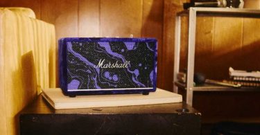 Enceinte Bluetooth Marshall Acton III en édition limitée Hendrix 60th Anniversary, posée sur un meuble dans une pièce aux murs lambrissés, recouverte de velours violet aux motifs cosmiques noir et violet, avec logo Marshall en argent et grille frontale assortie