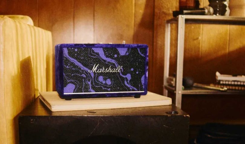 Enceinte Bluetooth Marshall Acton III en édition limitée Hendrix 60th Anniversary, posée sur un meuble dans une pièce aux murs lambrissés, recouverte de velours violet aux motifs cosmiques noir et violet, avec logo Marshall en argent et grille frontale assortie